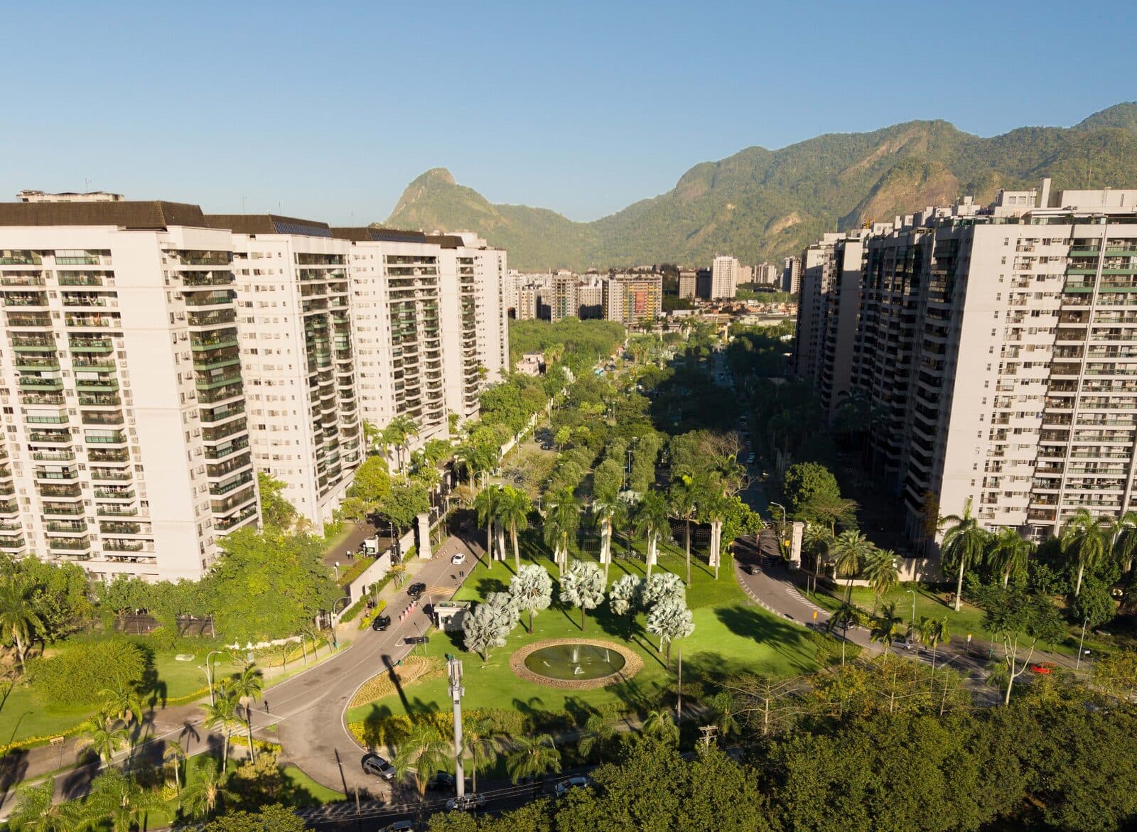 Vista aérea do Cidade Jardim, bairro planejado da Barra Olímpica, com praça, áreas verdes e montanhas ao fundo